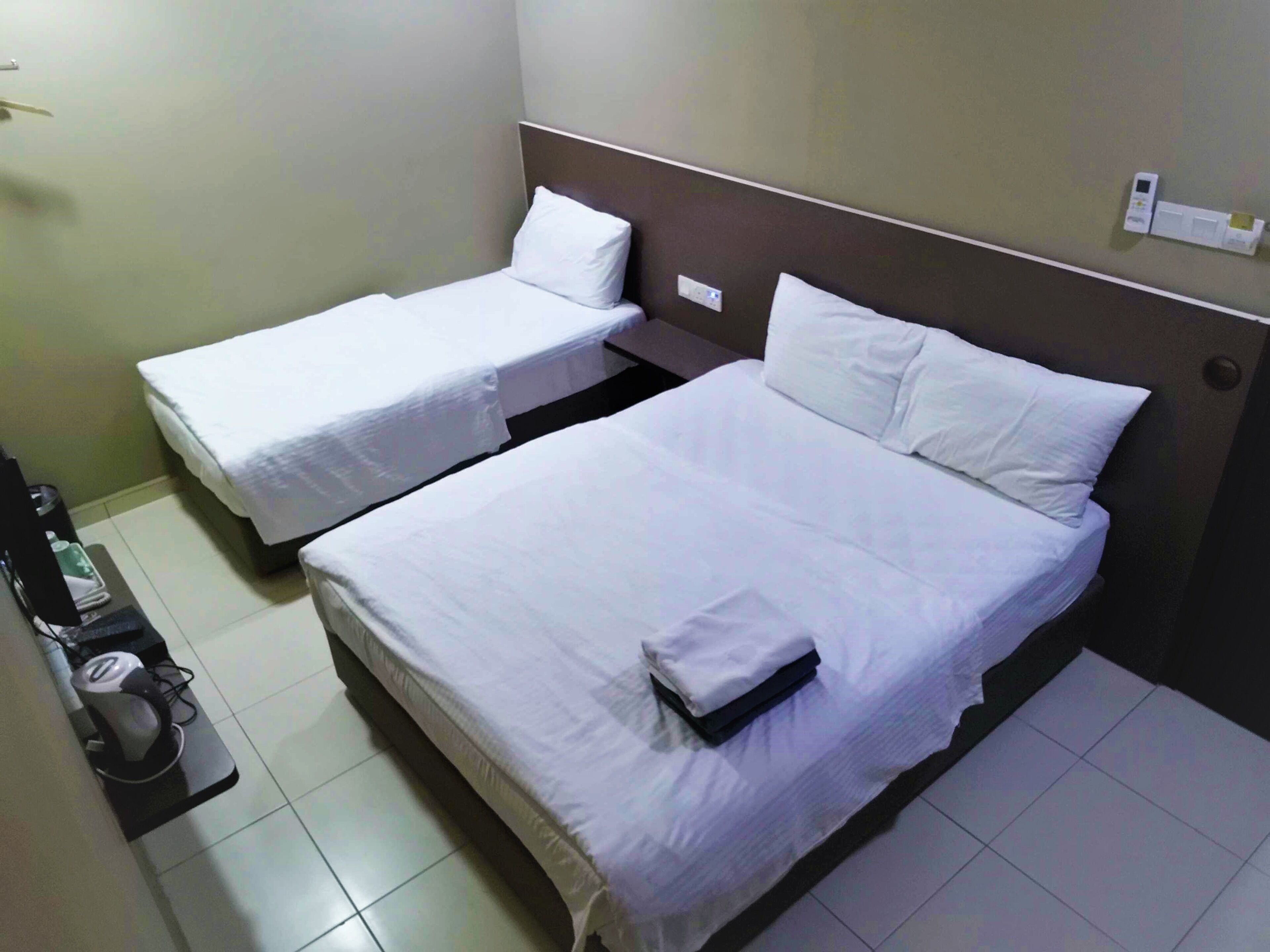 A hotel room with two single beds and a larger bed. There is a small table with a lamp and a bottle on it.