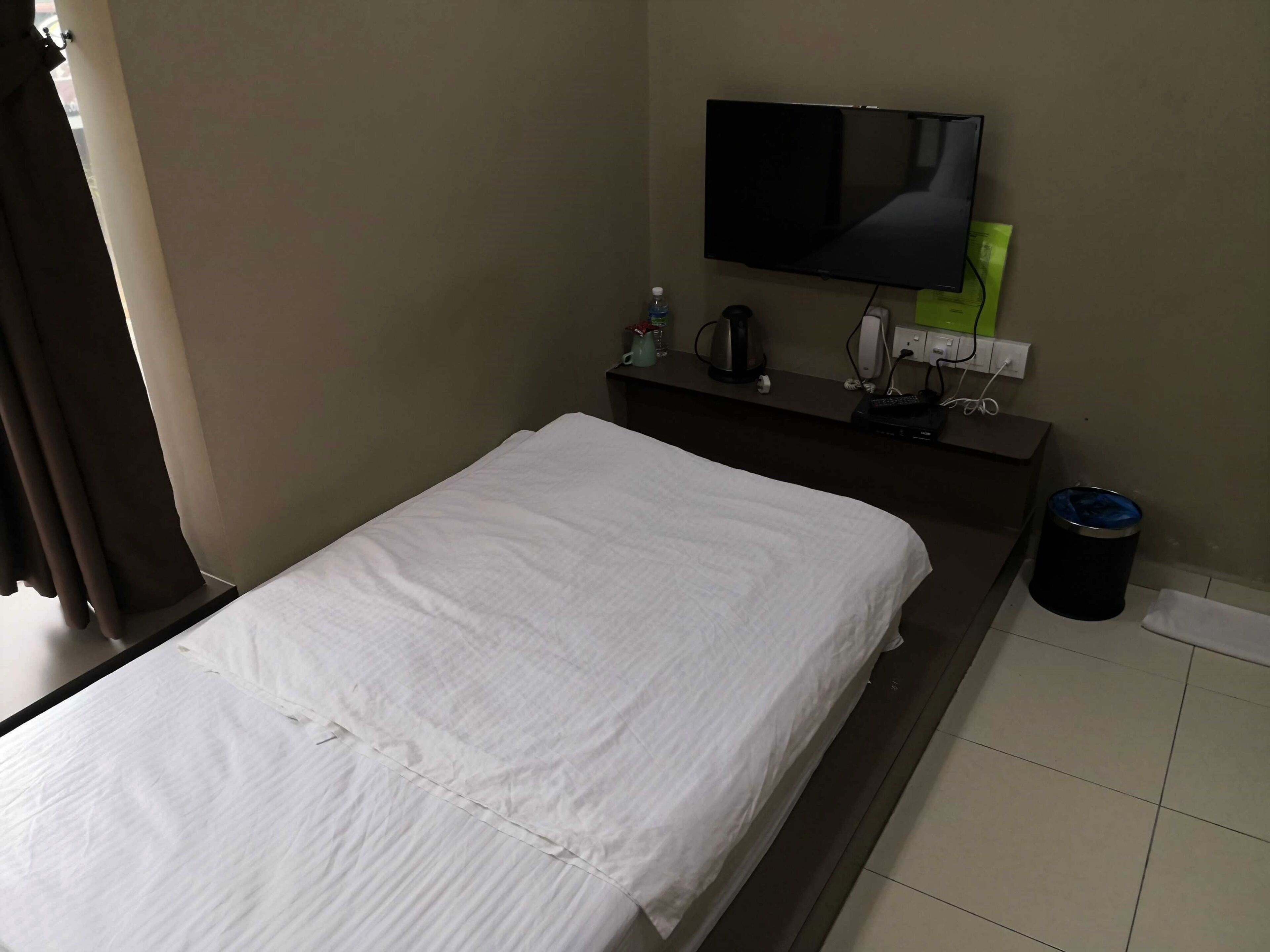 A hotel room with a bed, a TV mounted on the wall, a small table with a water bottle, a trash bin, and a power outlet.