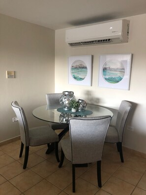 Un coin repas avec une table en verre entourée de quatre chaises grises, un climatiseur fixé au mur et deux œuvres d’art encadrées représentant des paysages.