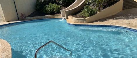 A swimming pool with a metal ladder, surrounded by a tiled deck and a landscaped area with bushes.