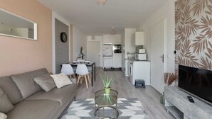 Una sala de estar moderna con un sofá, una mesa de centro y un televisor. Se observa una cocina al fondo con una lavadora y un comedor con sillas.
