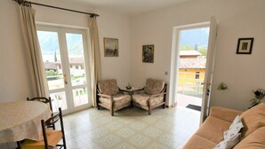 A living room with a patterned floor, a sofa, a dining table with chairs, and two armchairs. There are two doors leading outside, and the room has a view of buildings and greenery. - Lovely apartment for 6 people with WIFI, TV, balcony and pets allowed (Crone)