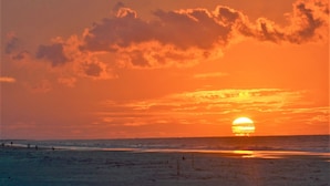 Escena de playa con atardecer, océano y gente a lo lejos.