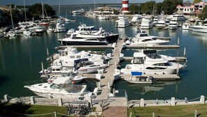 Un puerto deportivo con varias embarcaciones amarradas en muelles, un faro al fondo y una zona verde con bicicletas.