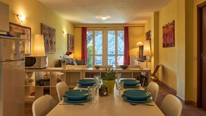 A dining area with a white table set for four, blue plates, and a vase with flowers. There is a microwave and a refrigerator in the kitchen area to the left.