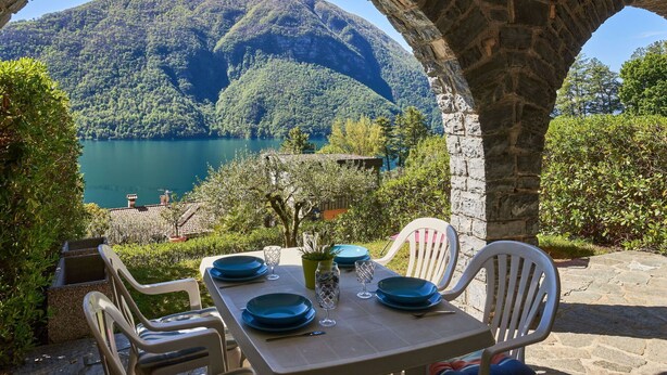 A table set for two with blue plates and glasses, overlooking a lake and mountains.
