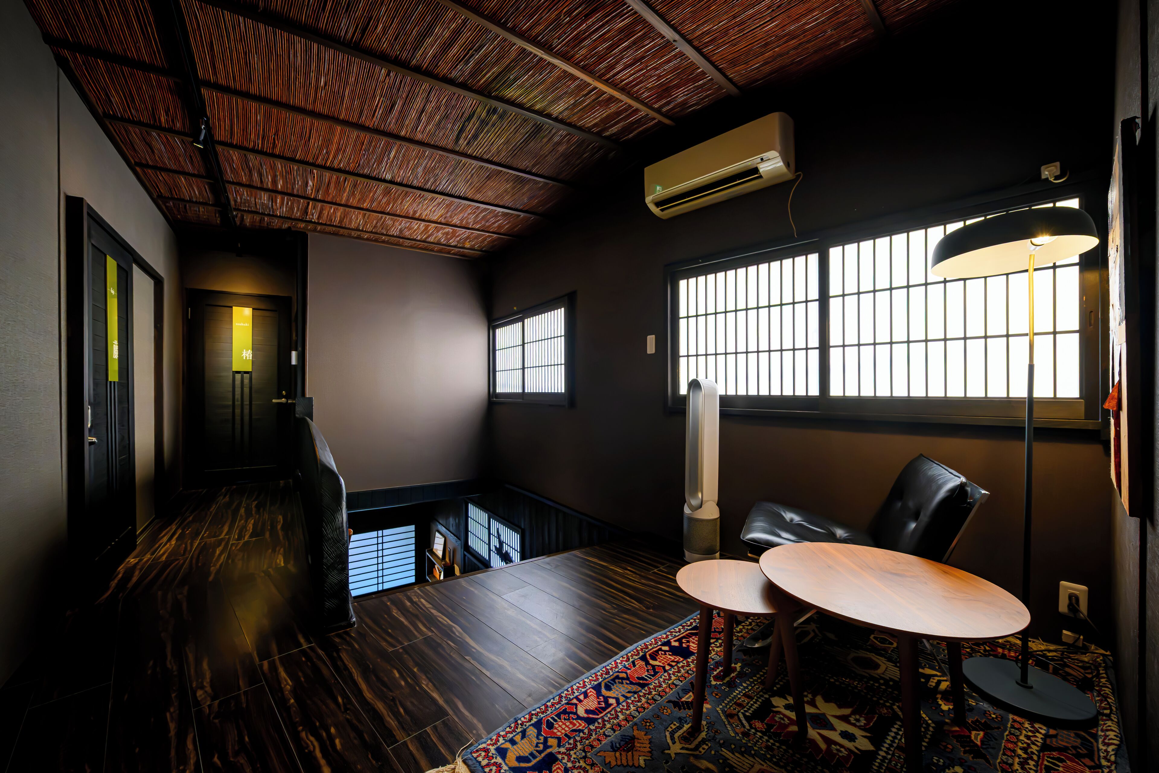 Een kamer met een hoge plafond, een houten vloer, een groot raam met jaloezieën, een tafel met stoelen, en een airconditioning.
