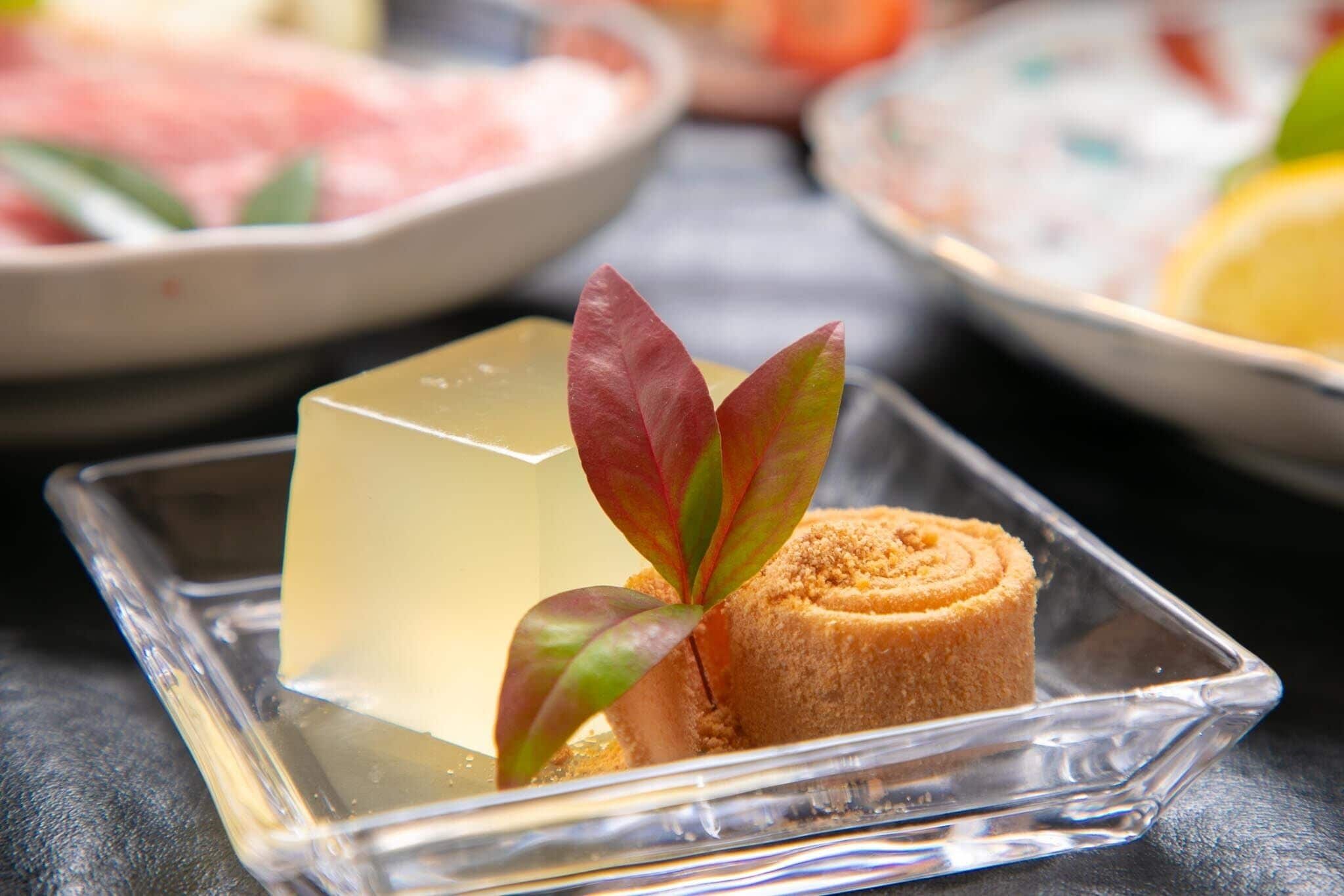 A clear glass dish with a cube-shaped jelly dessert and a rolled pastry, accompanied by a decorative leaf.