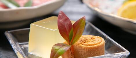 A clear glass dish with a cube-shaped jelly dessert and a rolled pastry, accompanied by a decorative leaf.