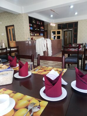 Un comedor con mesas dispuestas para una comida, una barra al fondo y un perchero con una chaqueta blanca.