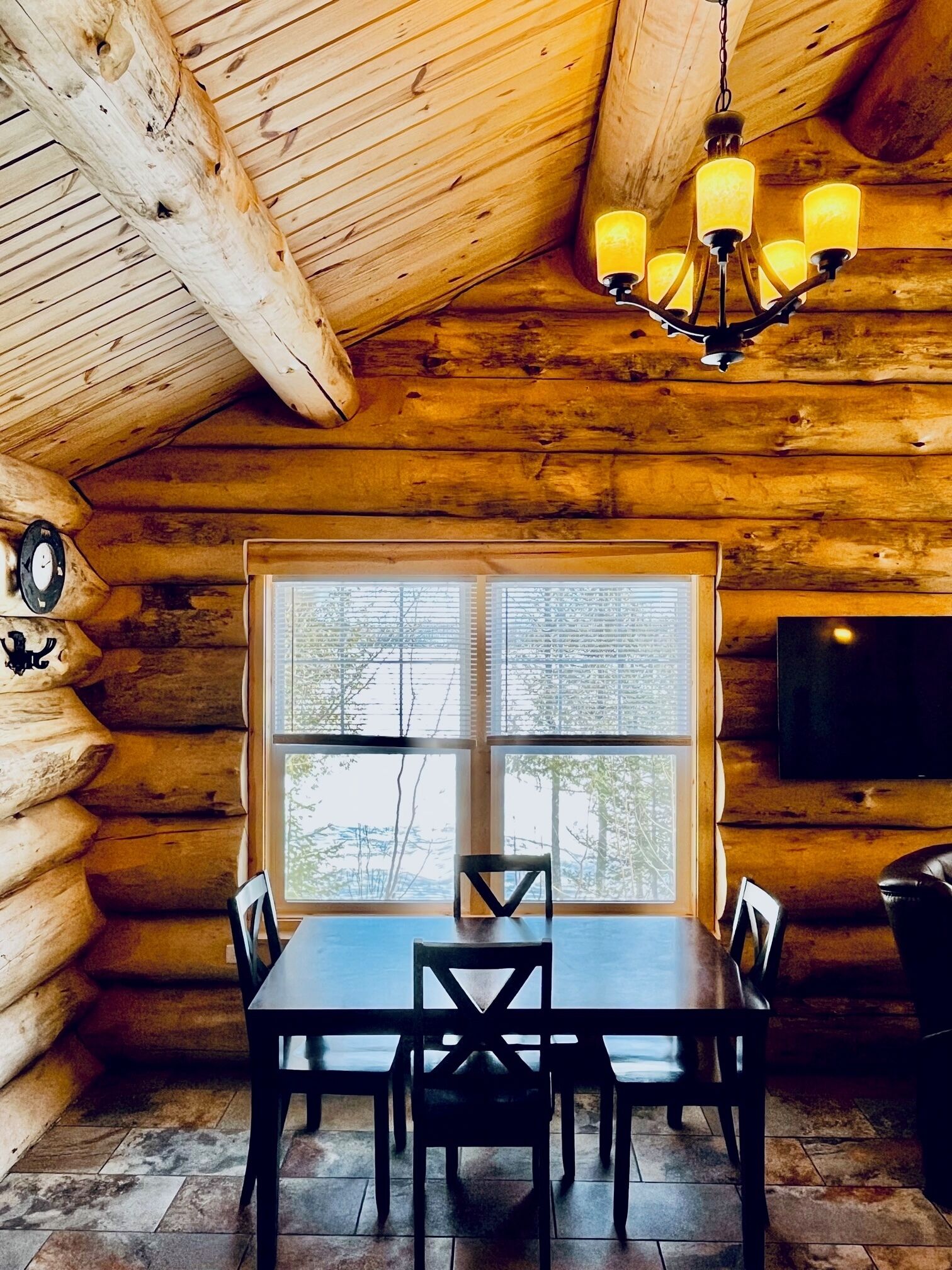 A log cabin interior with a dining table, chairs, a chandelier, a large window, and a television.