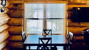 A log cabin interior with a dining table, chairs, a chandelier, a large window, and a television.
