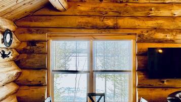 A log cabin interior with a dining table, chairs, a chandelier, a large window, and a television.