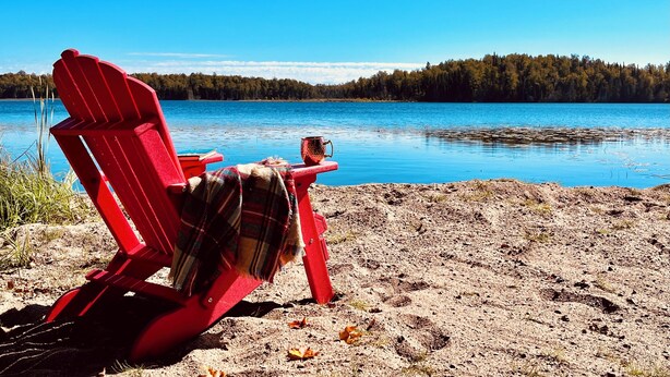A red Adirondack chair on a sandy beach with a view of a calm lake and forested shoreline.
