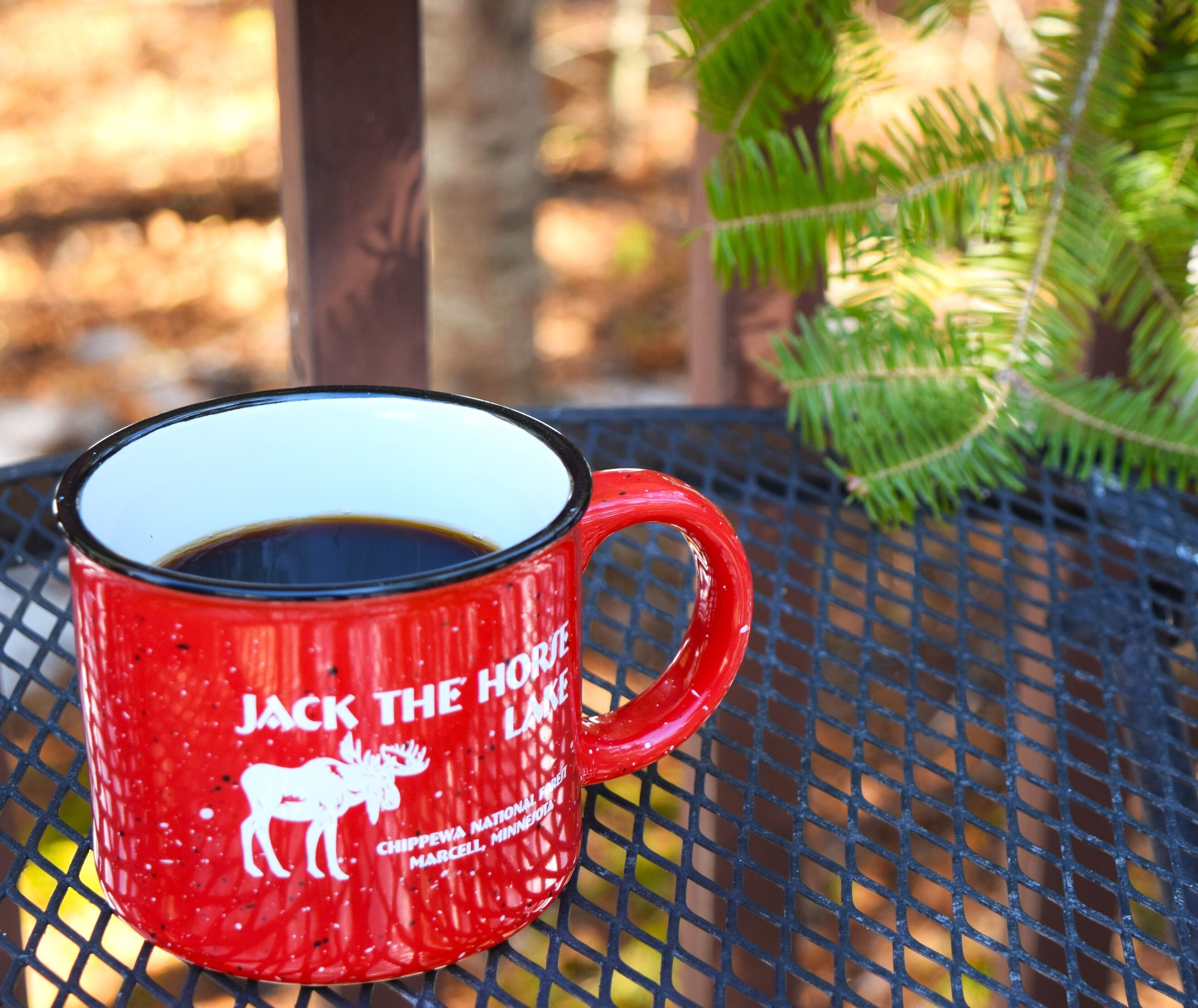 Una taza roja con texto blanco y la silueta de un caballo, sobre una mesa de metal al aire libre.