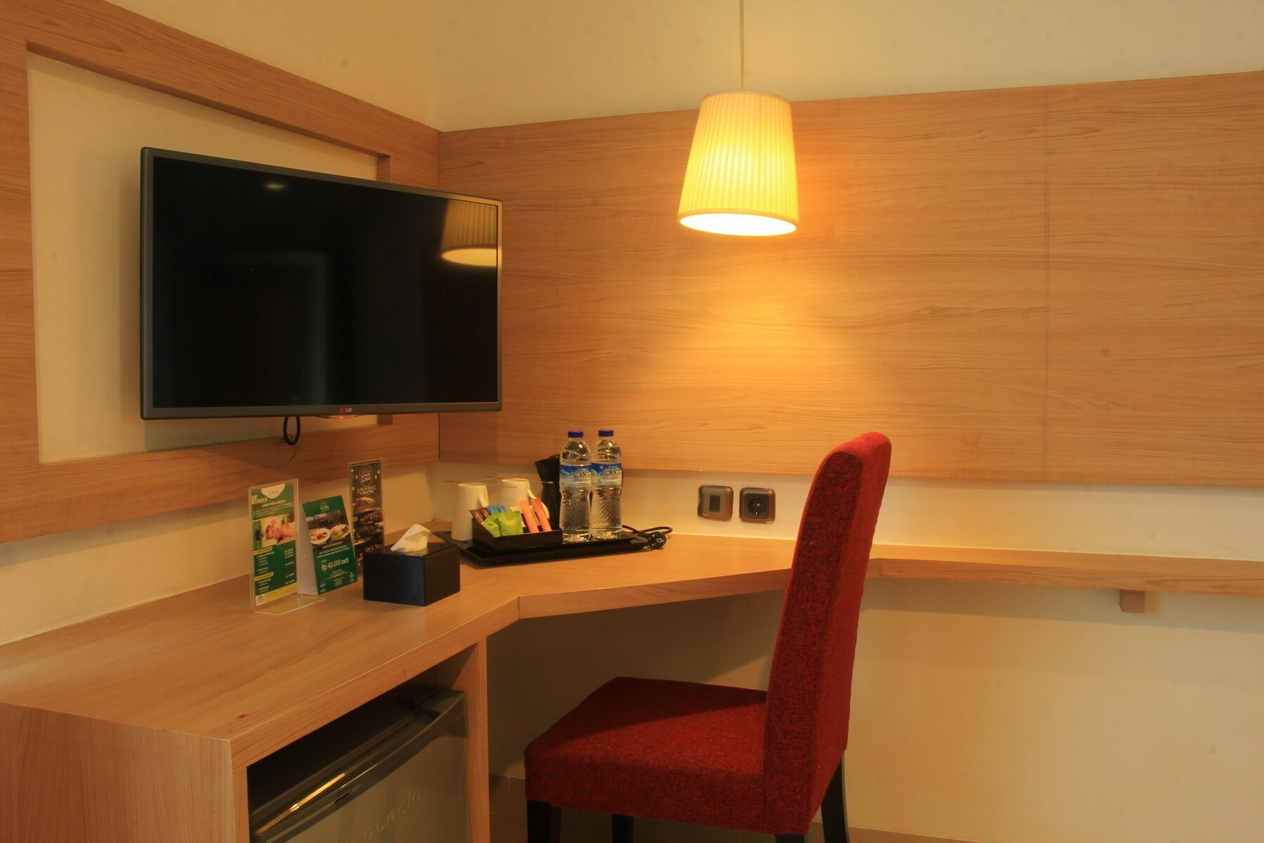 Quarto de hotel com escrivaninha de madeira, televisão, uma cadeira vermelha e um abajur.