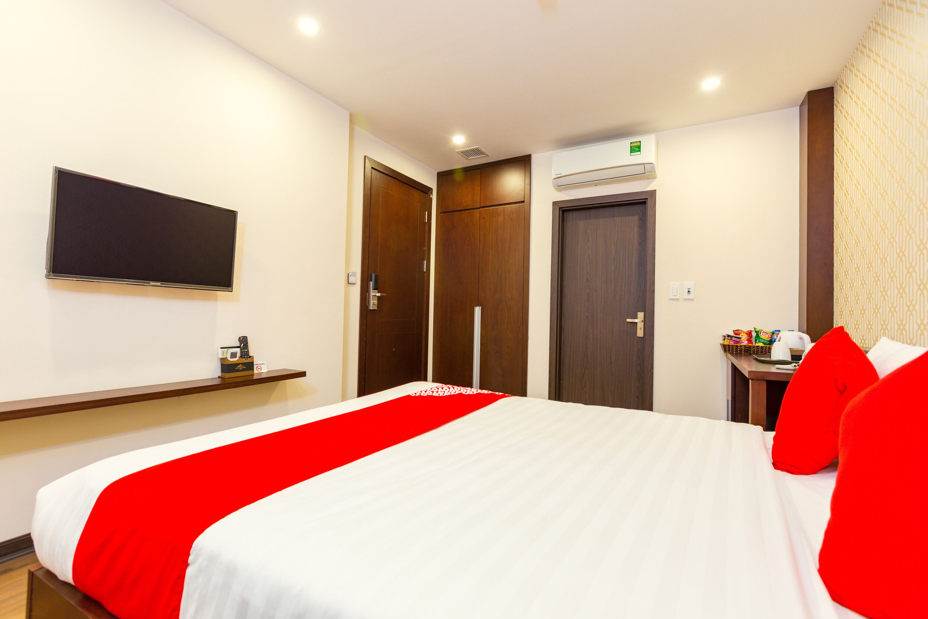 A hotel room with a bed, a TV mounted on the wall, a wardrobe, and a small table with a kettle and snacks.