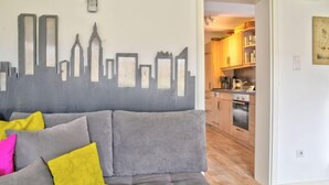 A living room with a grey sofa, a cityscape mural, a clock, and a kitchen visible through an open doorway. - Phileas by Interhome (Schwalenberg)