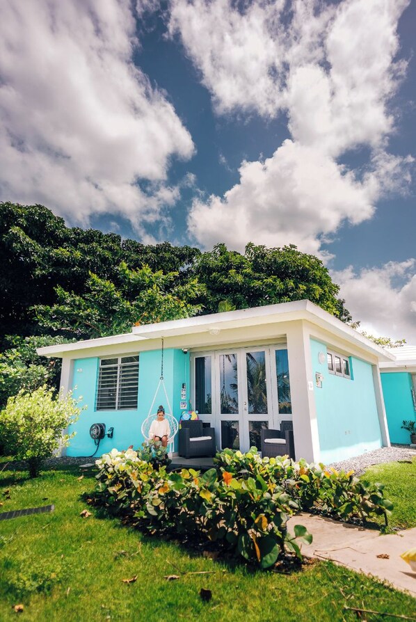 Une maison turquoise avec une véranda, un fauteuil suspendu et un petit jardin.