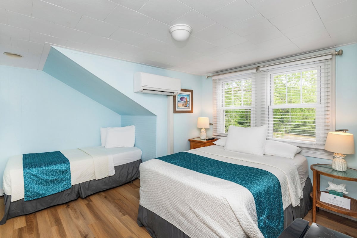 Une chambre d’hôtel avec deux lits, un escalier, un appareil de climatisation, une fenêtre avec des stores et une table de chevet avec une lampe.