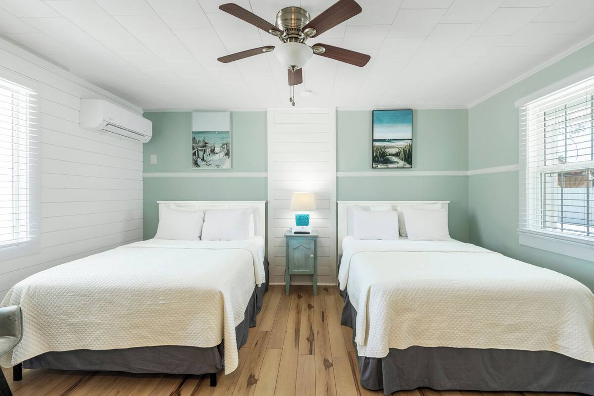 Une chambre avec deux lits, un ventilateur de plafond, une table de chevet avec une lampe et des œuvres d’art accrochées aux murs.