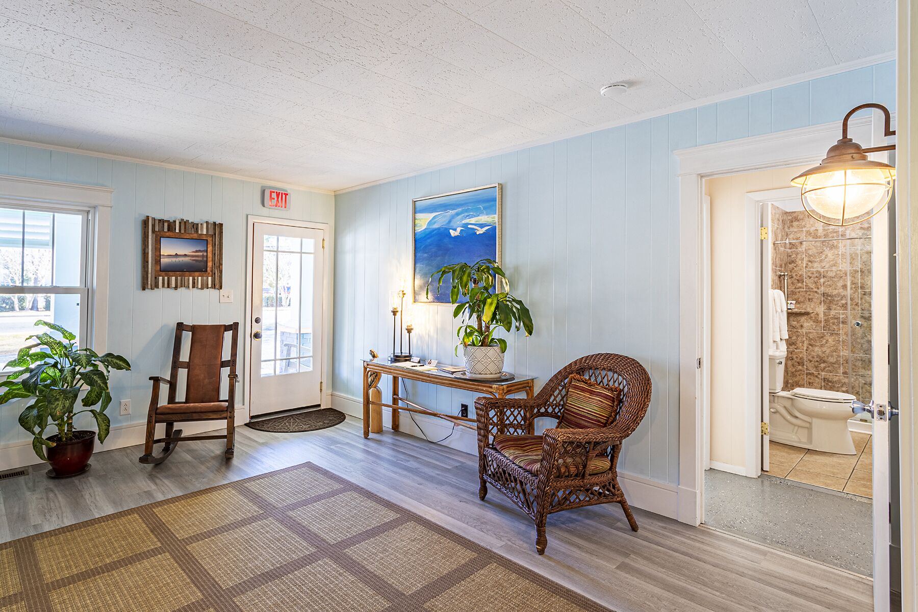 A room with a wooden chair, a table with a potted plant, a rug, and a painting on the wall.