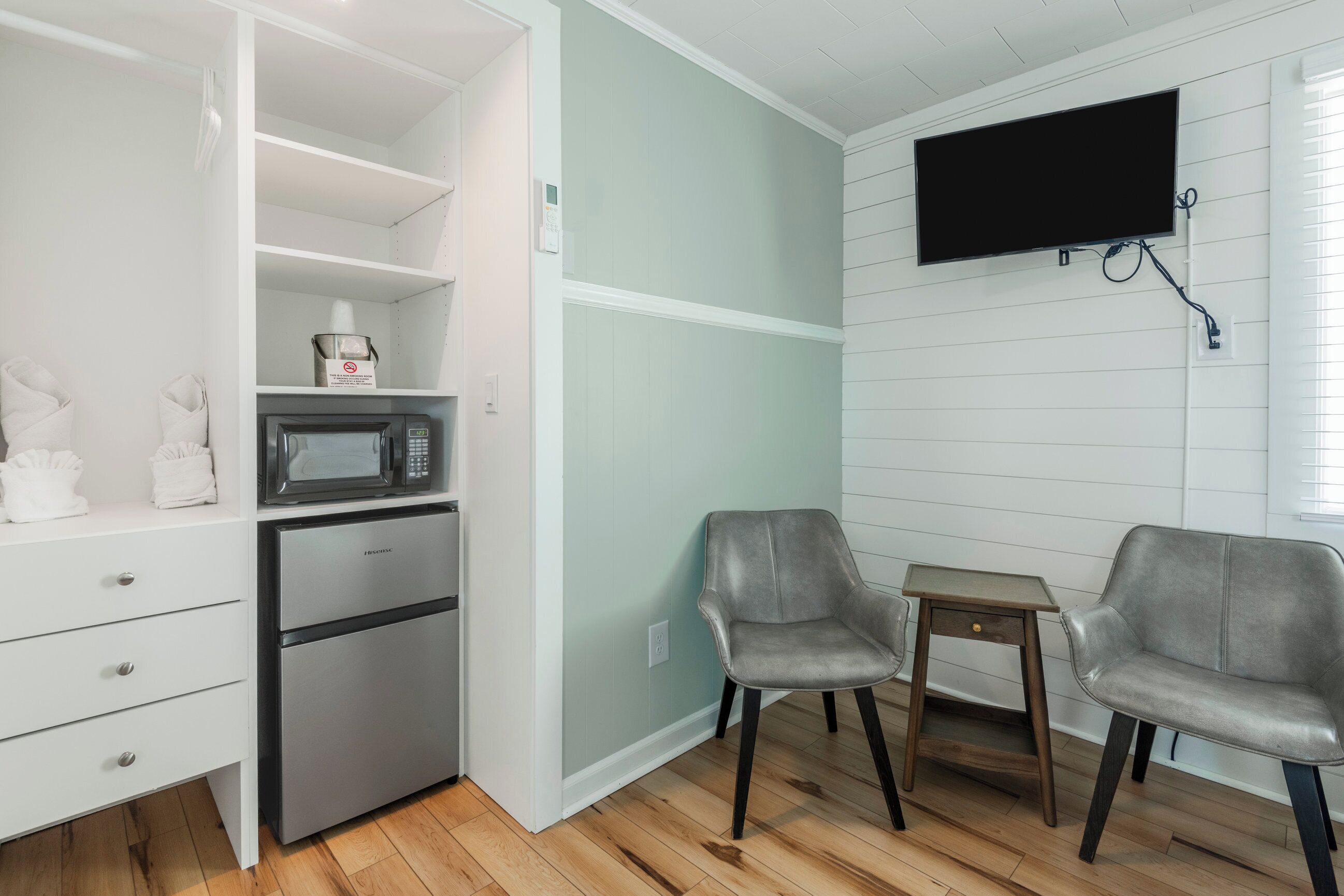 A compact kitchenette with a microwave, refrigerator, and storage shelves. There are two gray chairs and a small wooden table. A flat-screen TV is mounted on the wall.