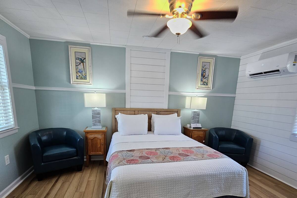 Une chambre à coucher avec un grand lit, deux tables de chevet, deux chaises, un ventilateur de plafond et des œuvres d’art accrochées aux murs.
