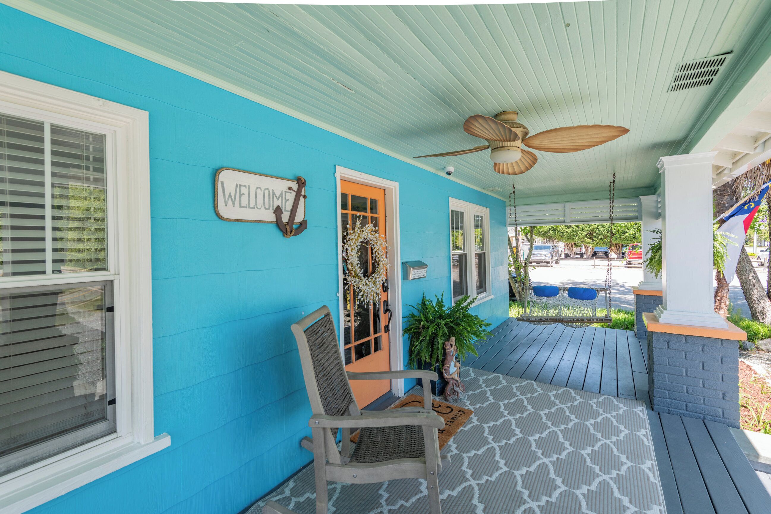 A porch with a blue exterior, a wooden chair, a welcome sign, and a door with an orange frame.