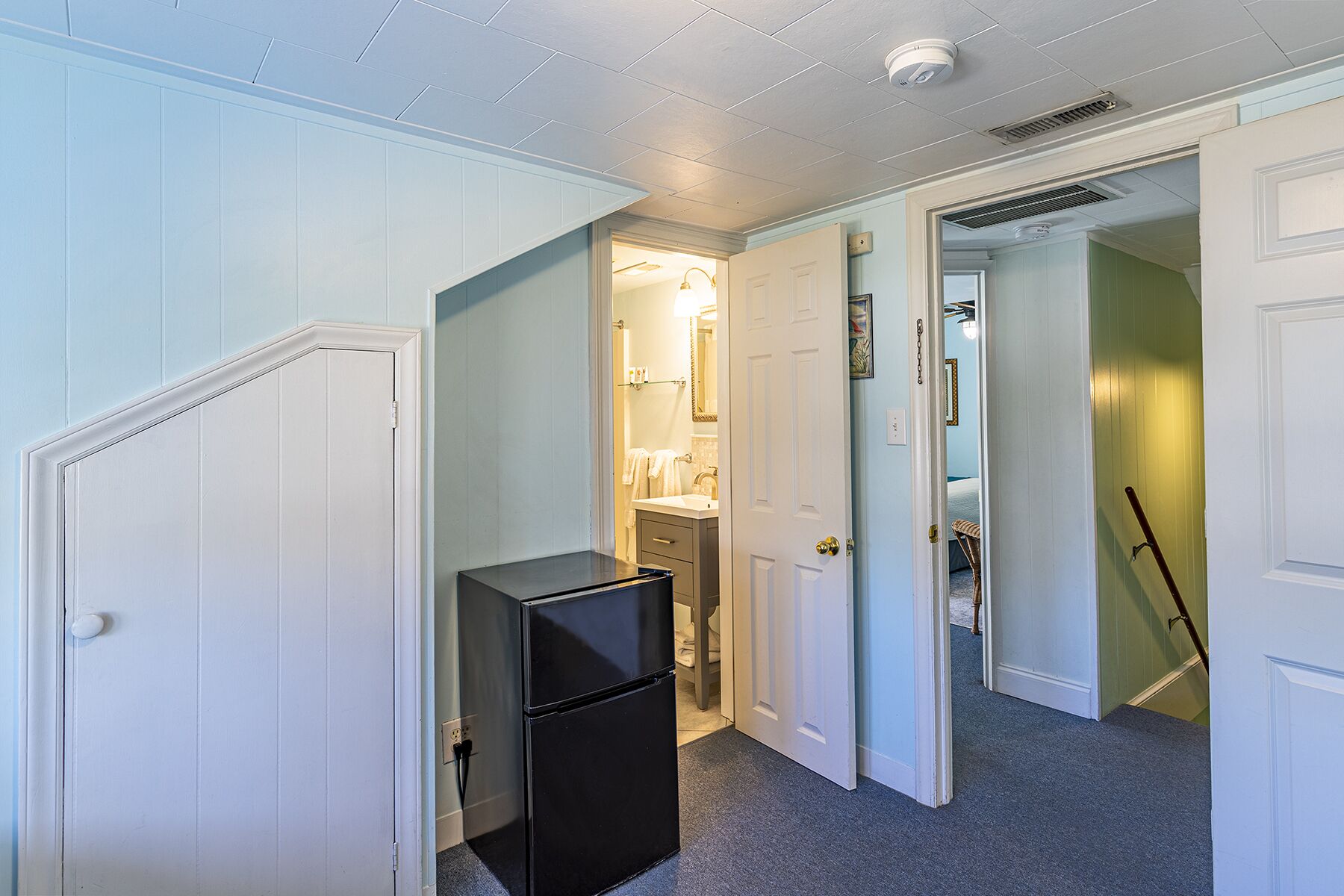 A hallway with a refrigerator, a bathroom with a sink and mirror, and a door leading to another room.
