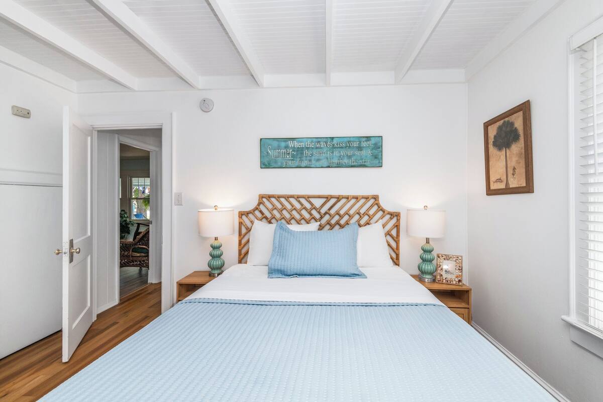 Une chambre à coucher avec un lit, une tête de lit en bois, un panneau décoratif fixé au mur, un tableau encadré et une fenêtre avec des stores.