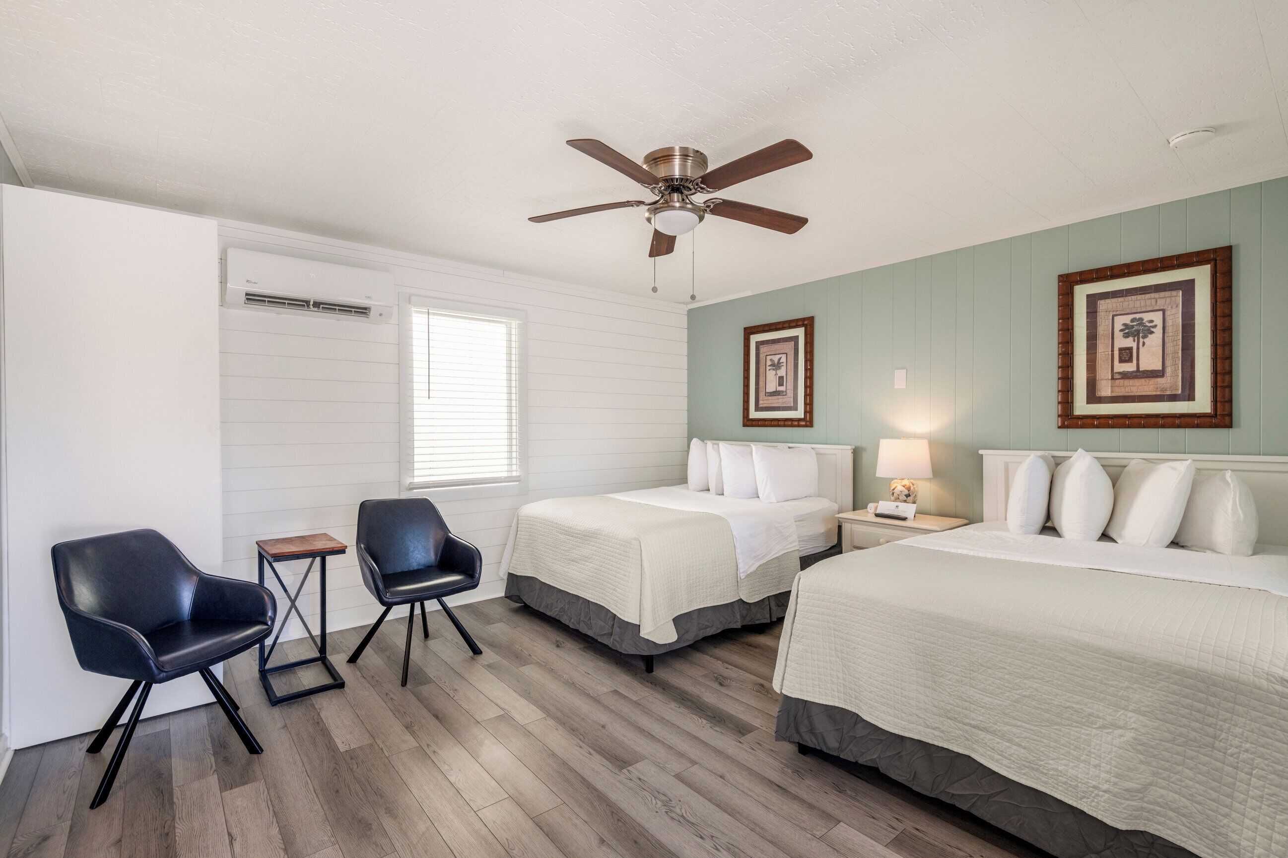 A hotel room with two beds, a ceiling fan, a small table, two chairs, and a window with blinds.