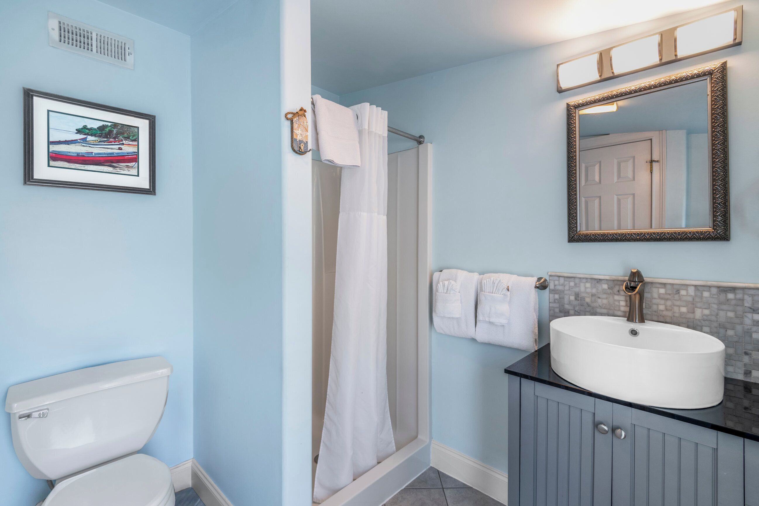 A bathroom with a white toilet, a shower with a white curtain, a sink with a mirror above it, and a framed picture on the wall.