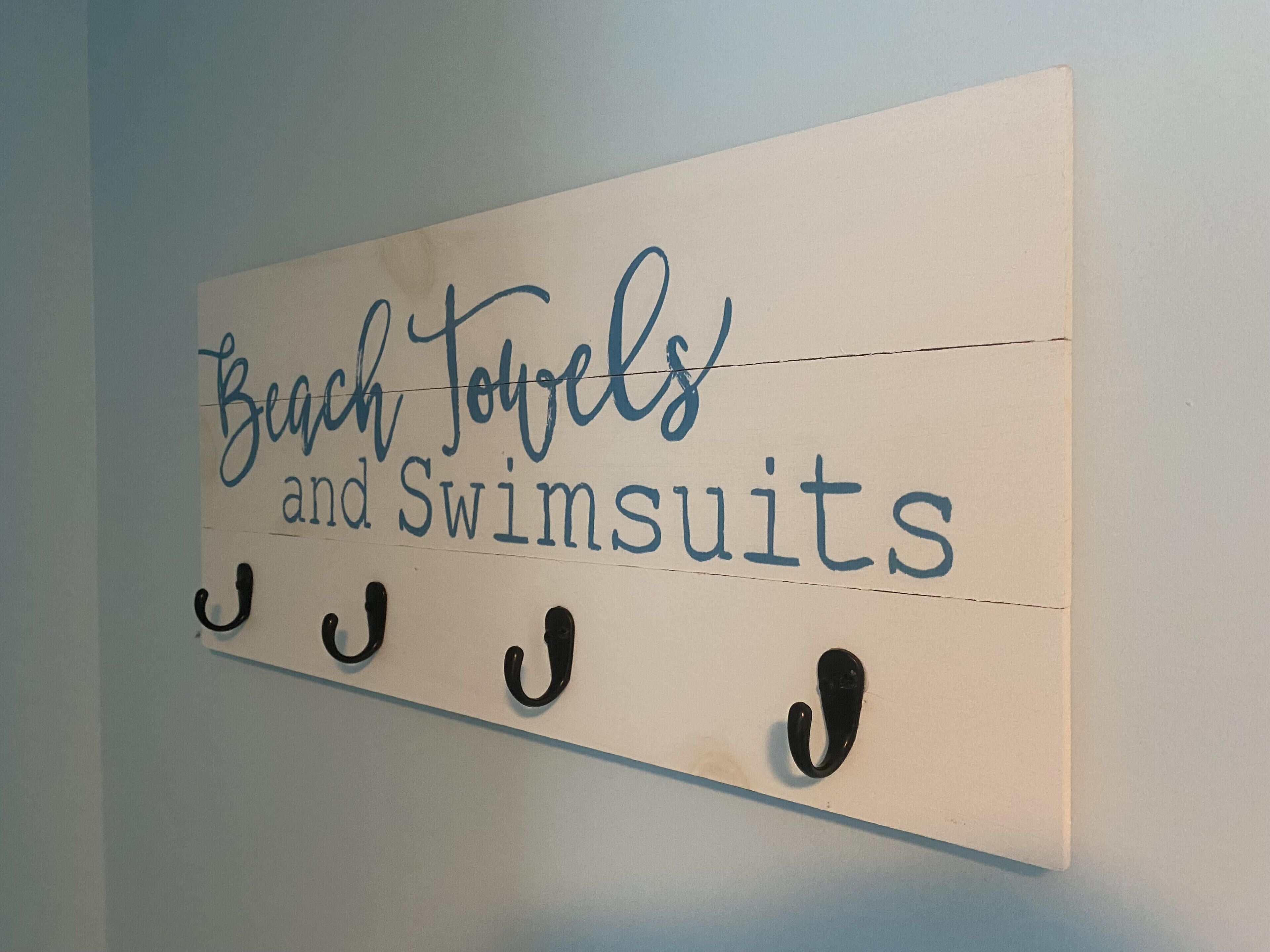 En skilt på væggen med teksten "Beach Towels and Swimsuits" og tre sorte kroge nedenunder.