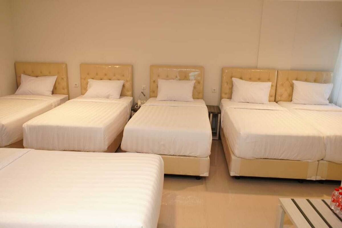 A hotel room with four single beds arranged in a row against a plain wall. Each bed has a headboard and a white mattress.