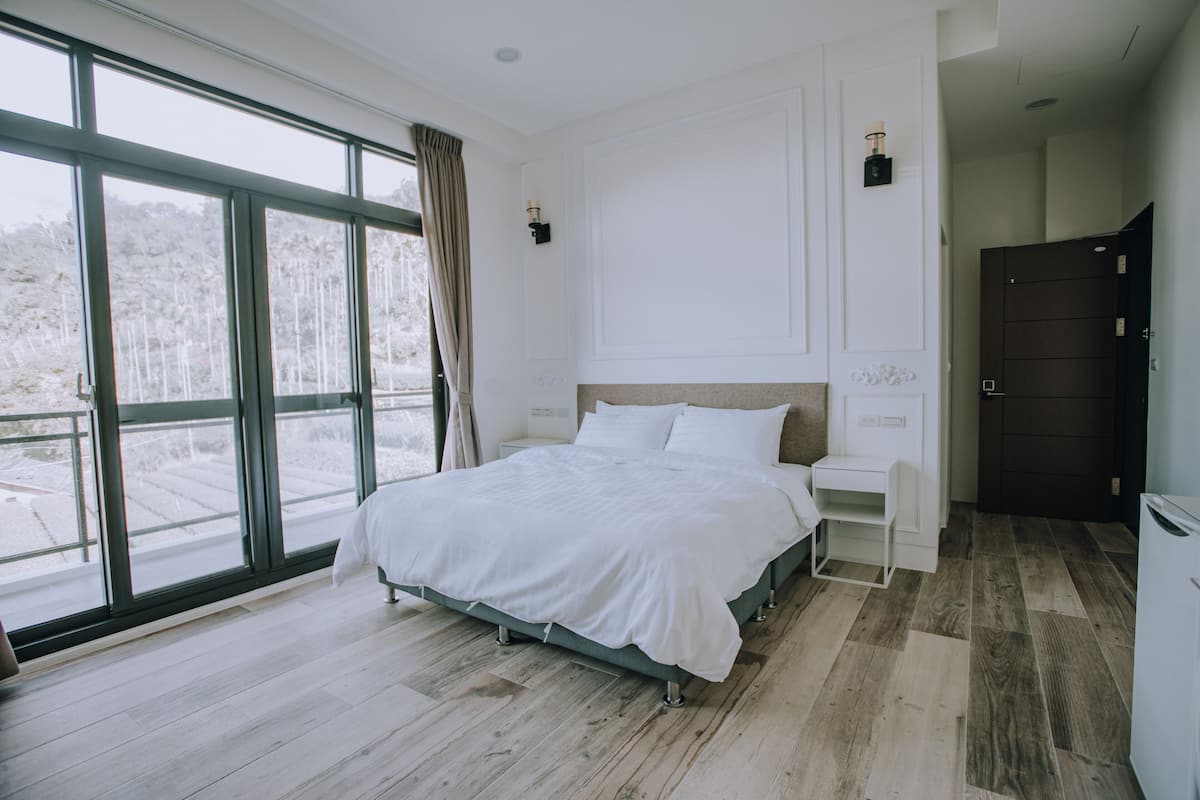 A bedroom with a large window, a bed, a nightstand, and a wardrobe.