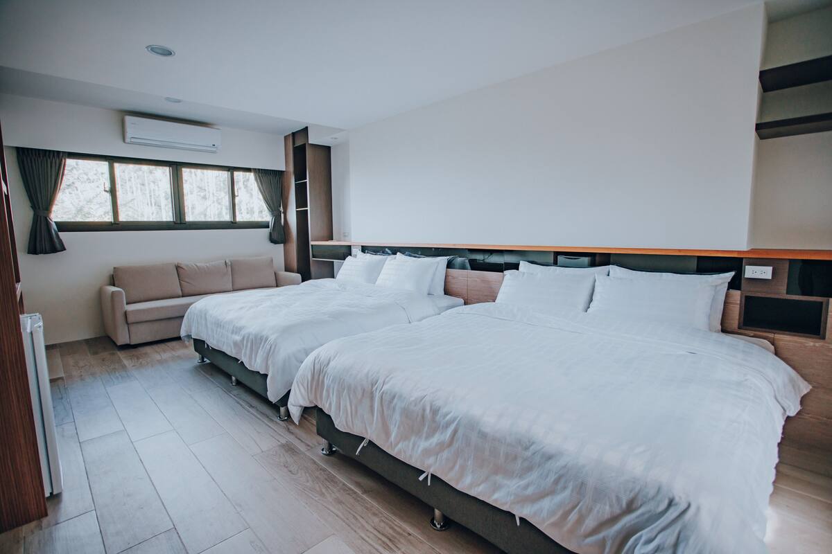 A modern hotel room with two beds, a sofa, a large window, and a built-in shelf.