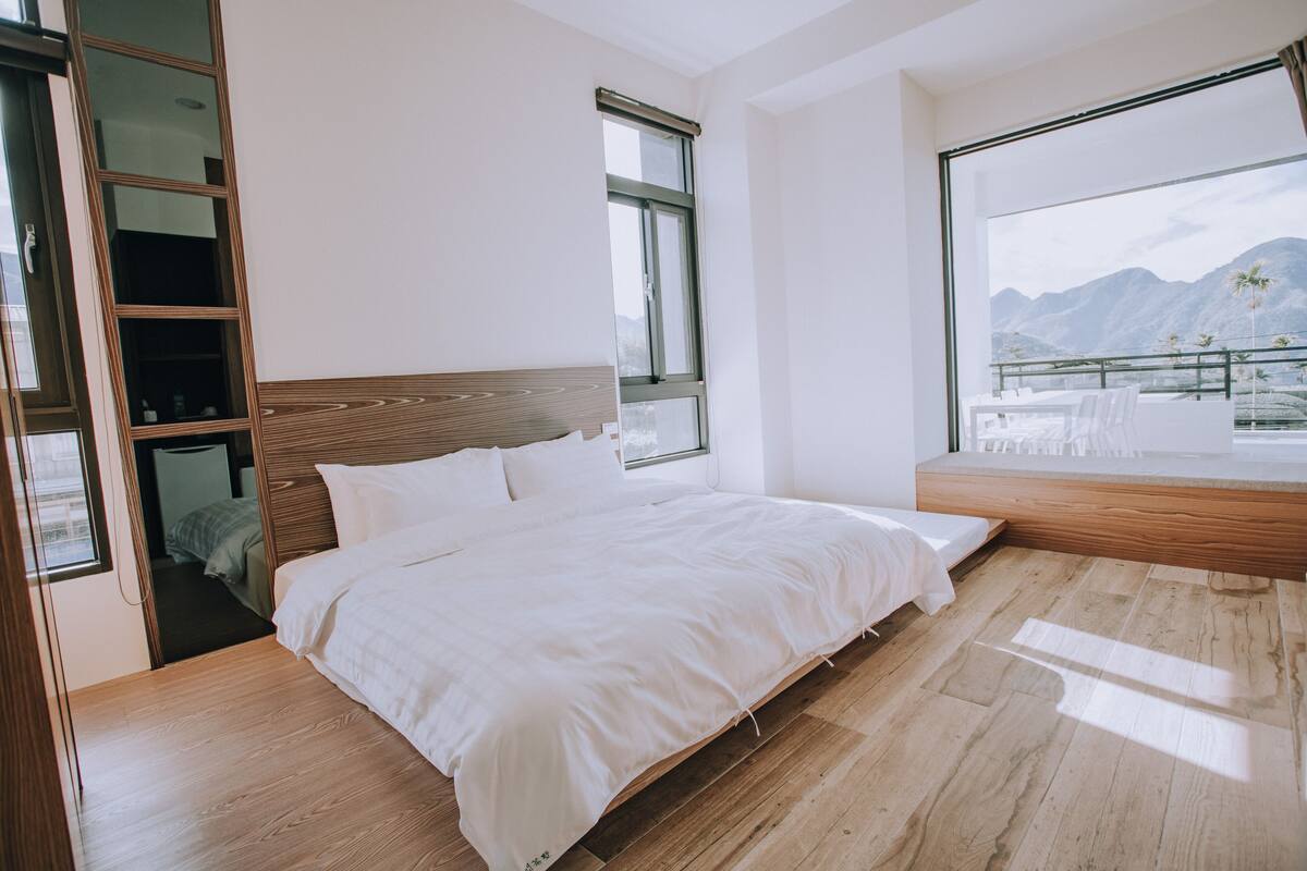 A modern bedroom with a wooden bed, large windows, and a balcony with a view of mountains.