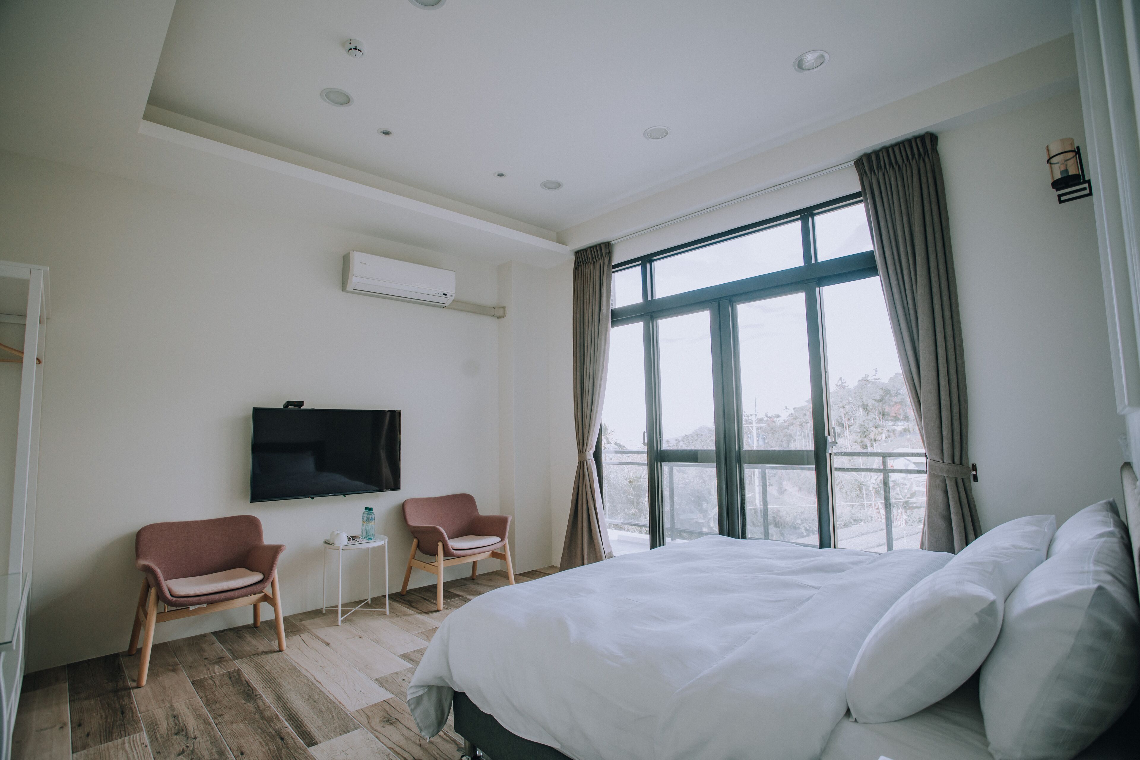 A hotel room with a large bed, two chairs, a TV, and a window with curtains.