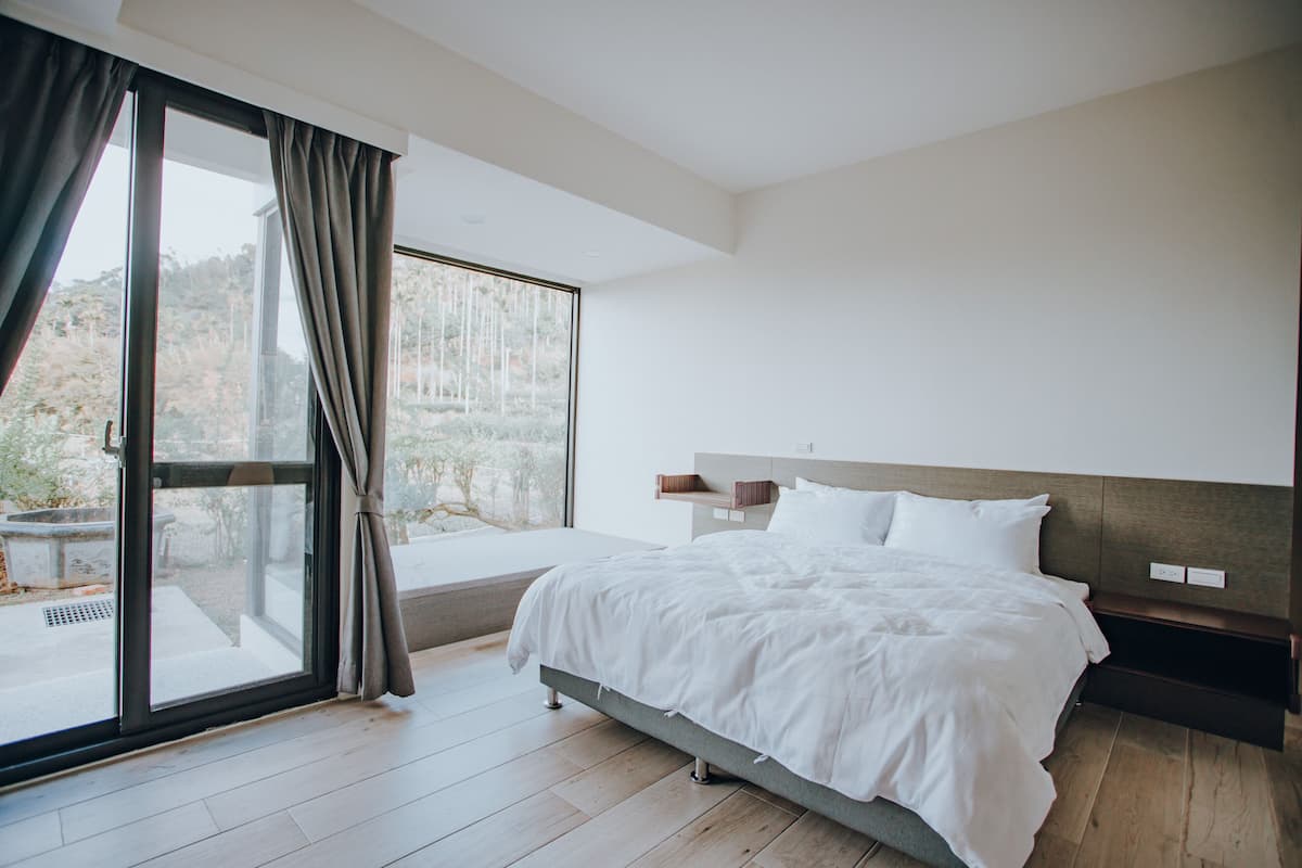 A bedroom with a large window, a bed, and a nightstand.