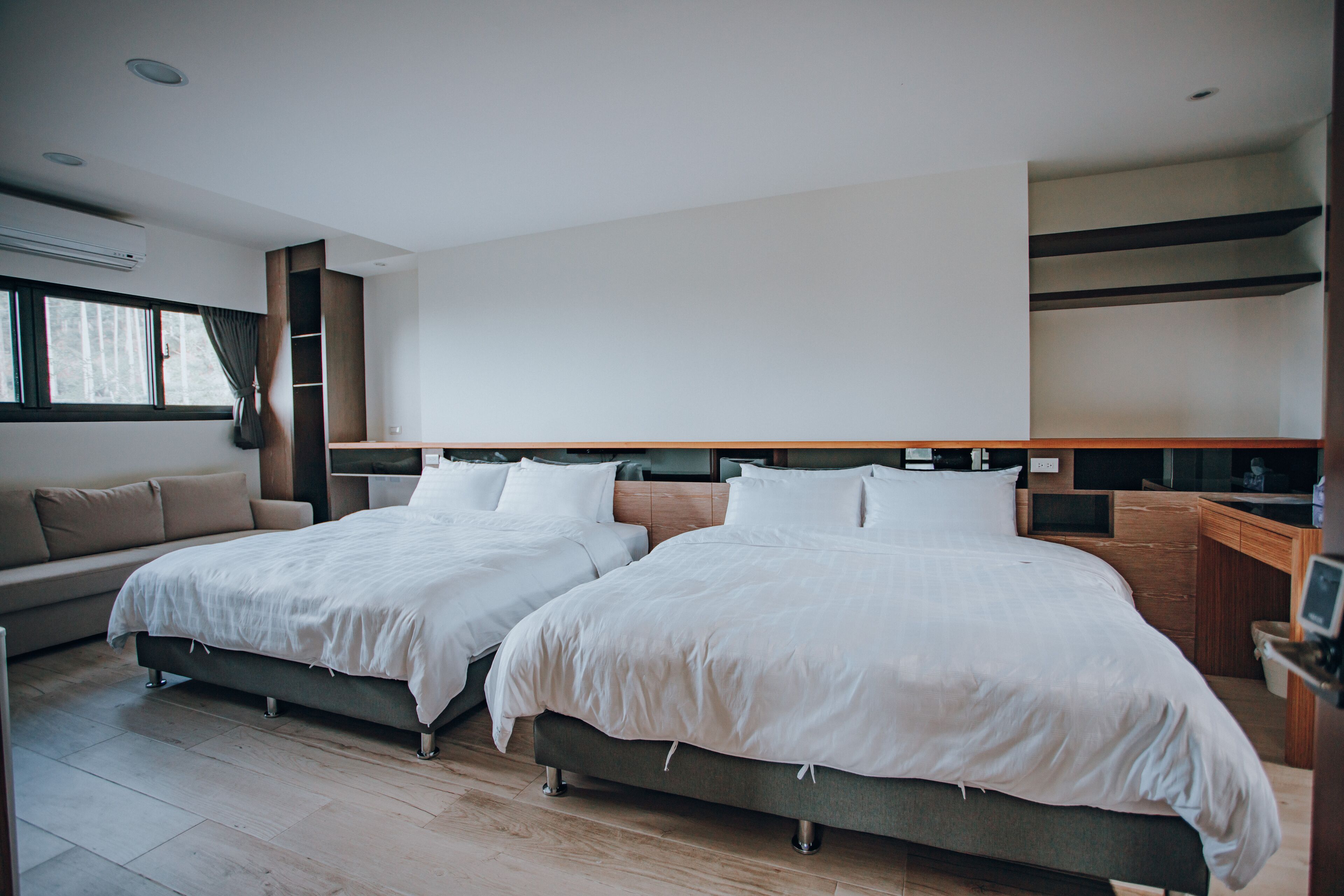 A modern hotel room with two beds, a sofa, a desk, and a large window.