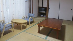 Une pièce de style japonais traditionnel, avec un sol recouvert de tatamis, une table basse, deux chaises, un téléviseur et des portes coulissantes.