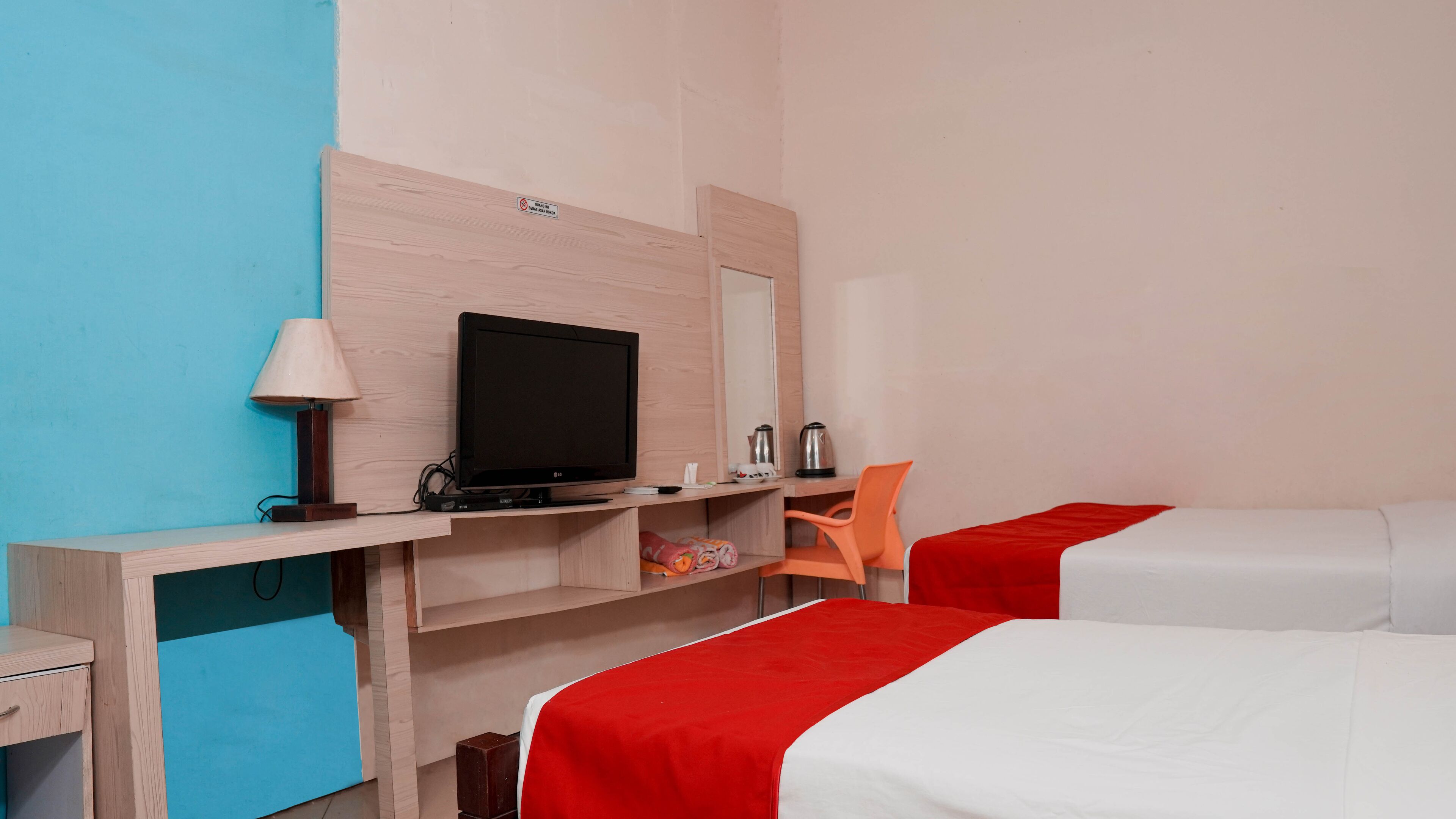A hotel room with a wooden desk, a television, a chair, and two beds with red and white bedding.
