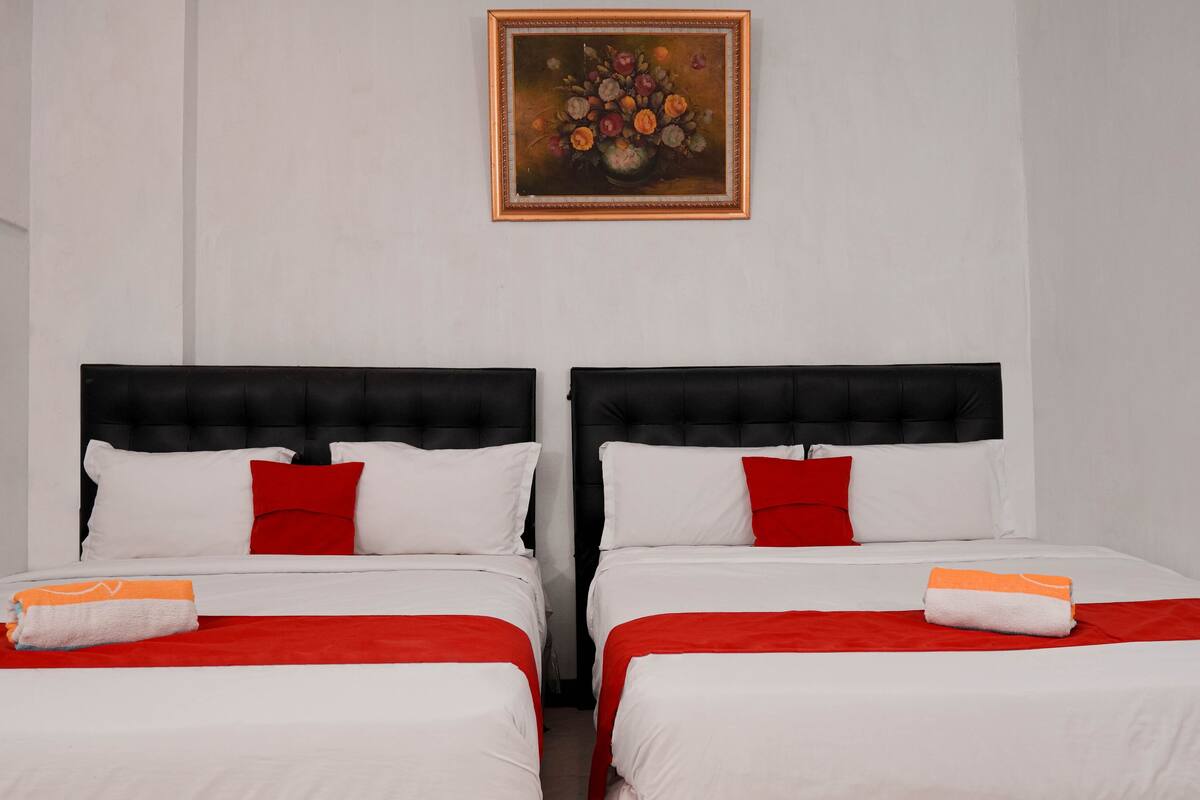 A hotel room with two beds, each with a black headboard, white bedding, and red pillows. A painting of flowers hangs on the wall above the beds.