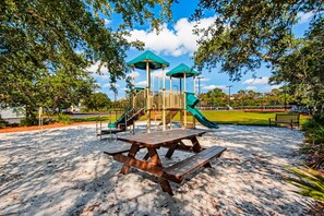 Playground area with a picnic table, slide, and climbing structure surrounded by trees and a bench.