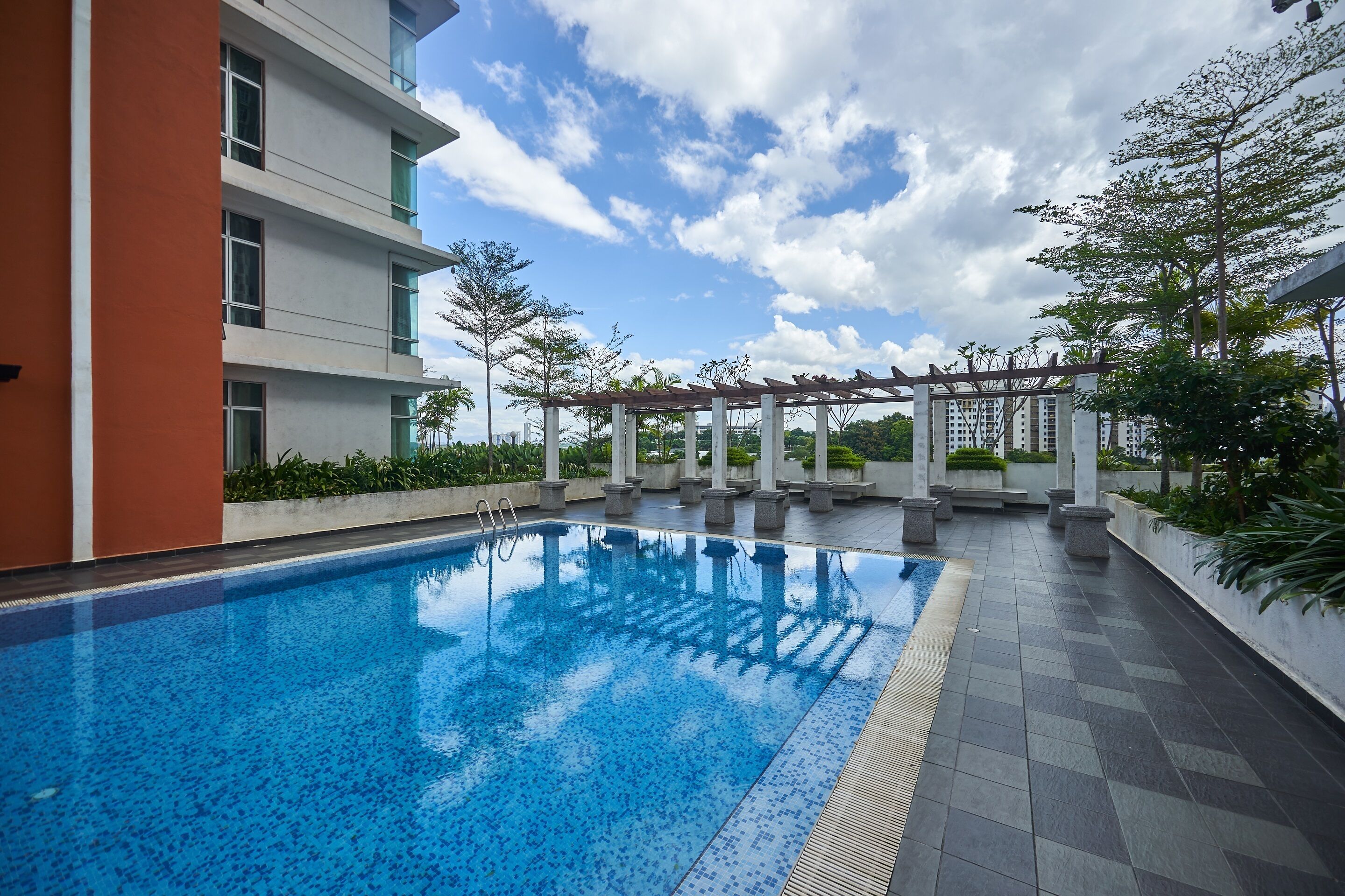 A swimming pool with a pergola structure, surrounded by a modern building with balconies and a clear sky.