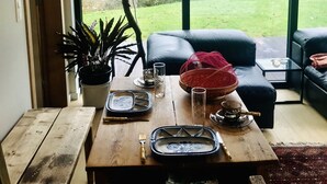 A cozy living room with a wooden table set for dining, a black leather sofa, and a large window offering a view of a grassy yard. - Panacée Guesthouses (Ronse)