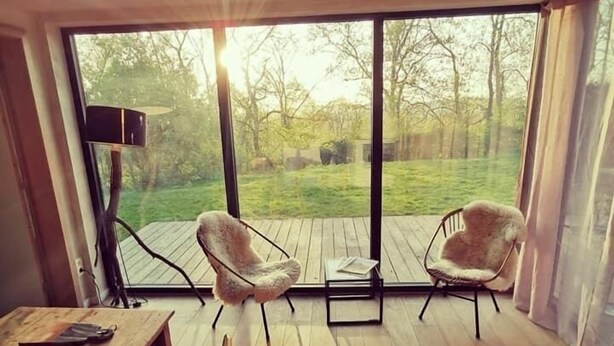 A room with a large sliding glass door leading to a deck, two upholstered chairs, a wooden table, and a floor lamp. - Panacée Guesthouses (Ronse)