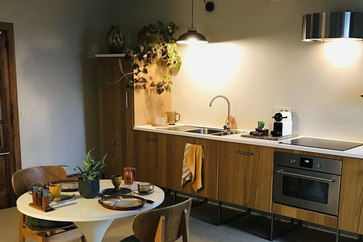 A modern kitchen with wooden cabinets, a stainless steel sink, and a built-in oven. There is a dining table with chairs and a decorative rug.