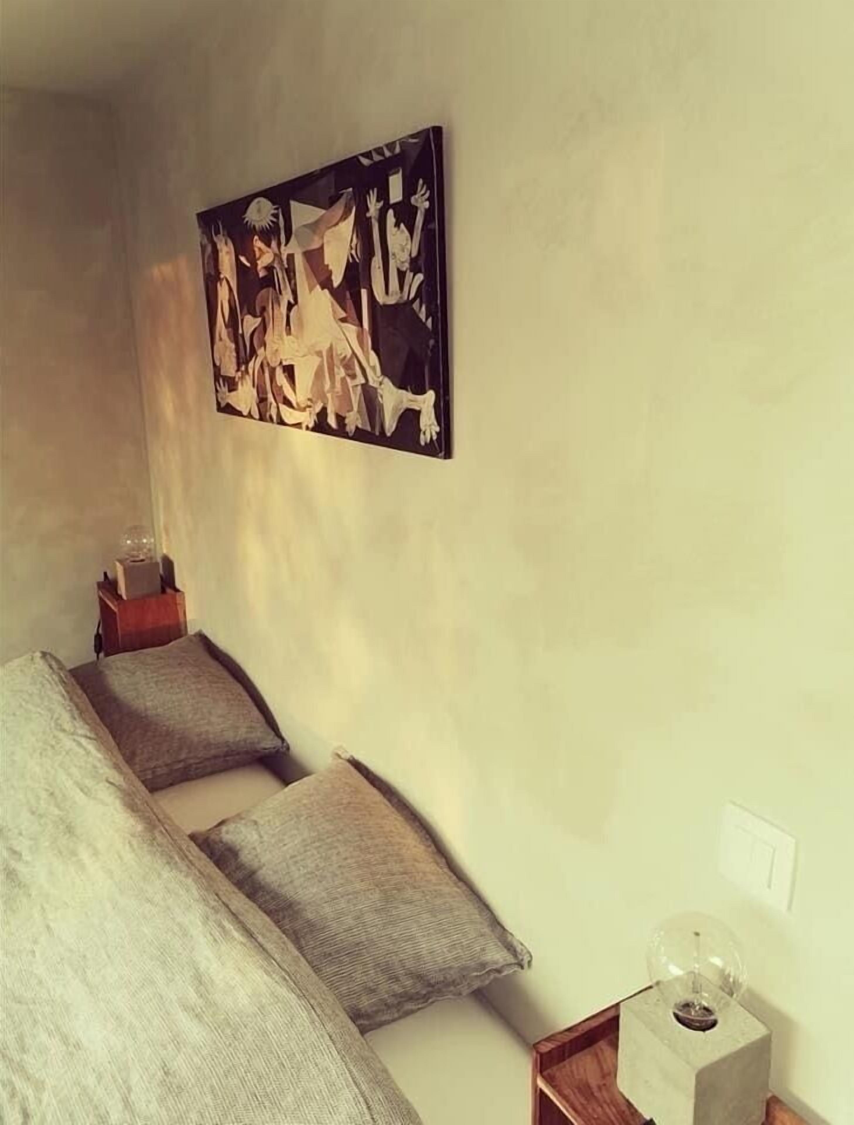 A bedroom with a bed, pillows, a nightstand, a lamp, and a wall-mounted abstract painting.